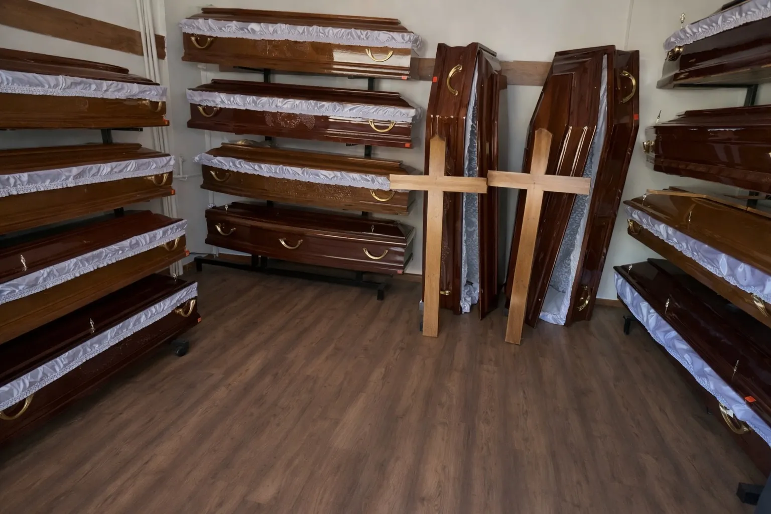 Mai multe sicrie din lemn sunt stivuite de-a lungul pereților unei încăperi cu podele din lemn. Două cruci de lemn sunt sprijinite în picioare printre sicrie, sugerând un magazin servicii funerare non stop sector 2 Bucuresti sau un showroom pentru nevoi funerare.